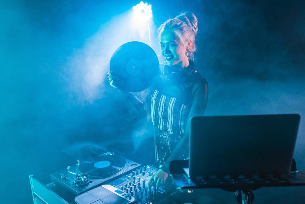 happy blonde dj girl standing near dj equipment and holding retro vinyl record in nightclub with